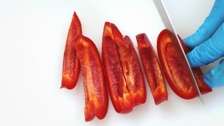 Sweet red pepper cutting close up on white kitchen board. Chef hands in blue gloves mince fresh papper with big iron knife. Cooking, preparing ingredients in professional restaurant. Top down viewの写真素材