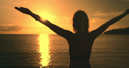 Close up woman face against bright orange sunset. Girl silhouette enjoys amazing colorful sunset, standing on a tropical beach, raises hands against orange dramatic sky and ocean waves. Holiday travelの写真素材