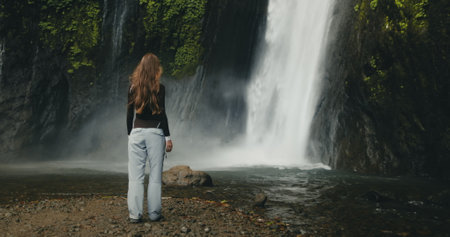 Tourist girl raising arms standing at powerful waterfall Air Terjun Munduk, Indonesia, Bali island. Woman posing, back view. Beautiful nature landscape. Travel, tourism, holidayの写真素材