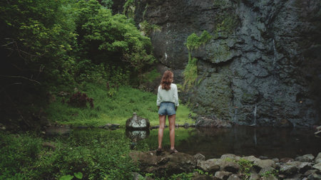 Tourist woman enjoy jungle waterfall landscape standing on rock stone. Girl relax outdoor lifestyle travel on summer holiday vacation. Picturesque scene panoramic shot. Dramatic dark toning, back viewの写真素材