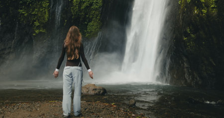 Tourist girl raising arms standing at powerful waterfall Air Terjun Munduk, Indonesia, Bali island. Woman posing, back view. Beautiful nature landscape. Travel, tourism, holidayの写真素材