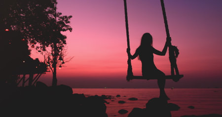 Woman silhouette sway on wooden rope swing against bright pink sunset. Vacation on beautiful seacoast. Travel, tourism, holiday on paradise Thailand Island. Nature landscape in sunrise.の写真素材