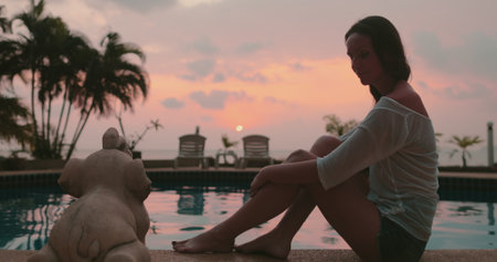 Woman relaxes sitting near pool on tropical island hotel resort. Bright colorful sunset sky and coconut palms reflected in water. Travel, tourism, holiday spa vacation.の写真素材