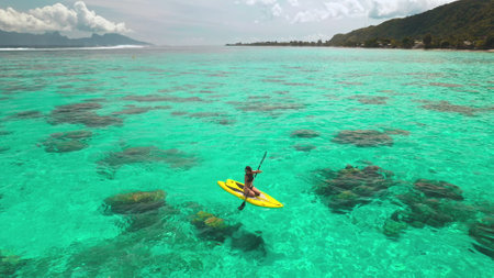 Aerial drone shot woman paddle boarding coral reef tropical island. Young female rows sitting on yellow paddleboard in crystal turquoise sea water. Outdoor lifestyle travel on summer holiday vacation.の写真素材