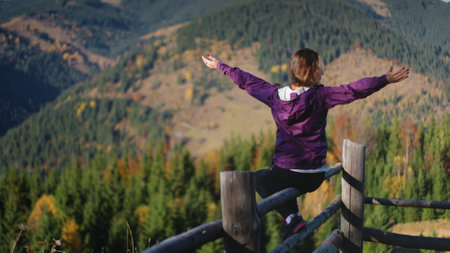 Woman enjoy mountain autumn morning landscape in Carpathians sitting on wooden fence. Raise hands against pine tree forest hill and meadow. Female tourist on summer vacation. Lifestyle holiday conceptの写真素材