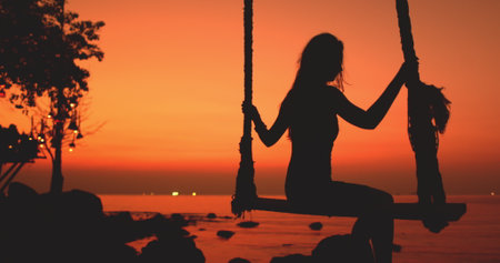 Romantic woman swing on sunset beach in Thailand. Girl silhouette against bright orange sky and sea water. Happy female relaxing, enjoy freedom outdoor travel on summer holiday vacation.の写真素材