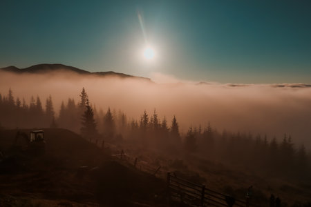 A misty morning in mountains, with sun peeking through clouds and casting a warm glow over the landscape. The silhouette of forest trees and hills are visible in the distance. Carpathians mountainsの写真素材