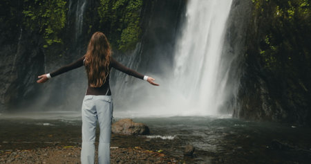 Tourist girl raising arms standing at powerful waterfall Air Terjun Munduk, Indonesia, Bali island. Woman posing, back view, . Beautiful nature landscape. Travel, tourism, holidayの写真素材