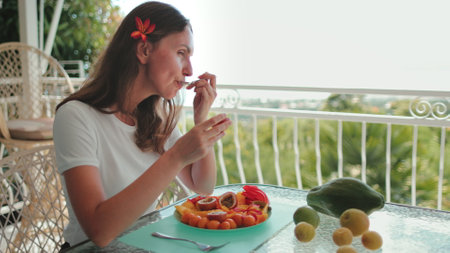 Woman eating tropical fruits sitting on the balcony with jungle view. Caucasian girl with flower in hair using a spoon try passion fruit, papaya, mango. Healthy eating, breakfast, organic vitamin foodの写真素材