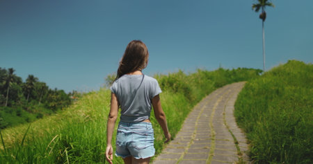 Girl walking green field asian Bali landscape in sunny day. Woman walking Campuhan Ridge Walk Trail, Indonesia, Ubud. Back view long hair tourist girl enjoy green hill nature landscape. Slowの写真素材