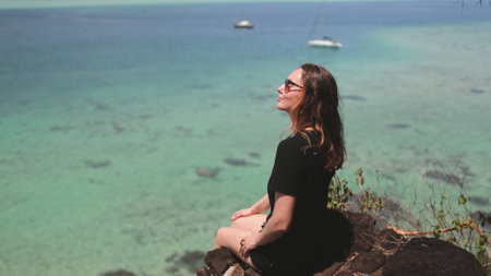A woman sits on top of a large rock near the ocean, with waves crashing in the background.の写真素材