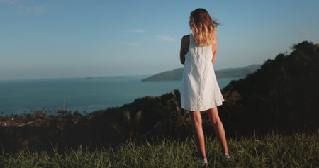 Woman in white dress stands on hill ocean and a small island in the distance. Girl relax tropical island , enjoy wild nature landscape. Outdoor lifestyle travel, summer holiday vacation,の写真素材