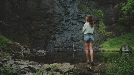 Woman enjoy jungle waterfall standing on rock. Travel to Bali island, Indonesia. Outdoor lifestyle travel on summer holiday vacation. Dark dramatic toning. Back viewの写真素材