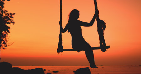 Woman relax enjoy swing ocean sunset sky. Female silhouette swinging tropical landscape, scenic orange sky, sea waves. Outdoor lifestyle travel on summer holiday vacation, freedom. Slow motionの写真素材
