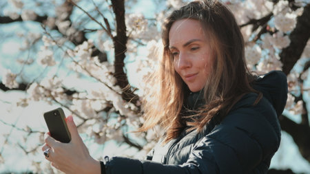 Girl making selfie with white sakura flowers in Tokyo, Japan. Woman enjoy spring blossom happy smiling looking to the camera. Sunny spring outdoors activity. Spring blossom of sakura tree in city parkの写真素材