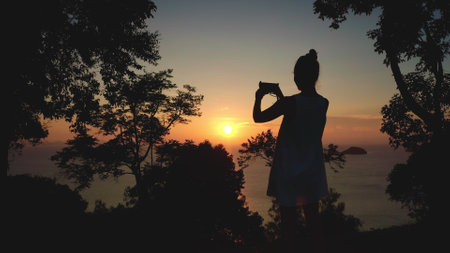 Woman enjoy sunset panorama sea view. Female silhouette standing in forest take photo on mobile phone. Bright orange sky sun in background. Exotic nature landscape. Travel, summer holiday relax, joyの写真素材