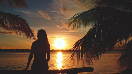 Silhouette of a young woman enjoying a beautiful sunset over the ocean, sitting on a wooden pontoon, framed by palm treesの写真素材