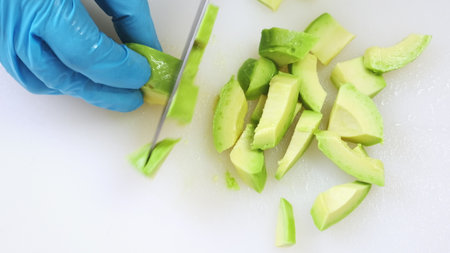 Half of green avocado cutting close up on white kitchen board. Chef hands in blue gloves chop fresh...