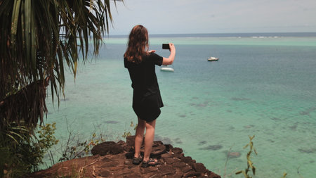 A woman standing on the shore, holding a camera, capturing a boat sailing in the vast ocean.