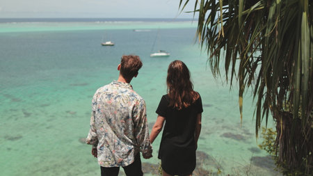 Young couple holding hands, admiring the ocean from a tropical cliff, with turquoise waters blending...