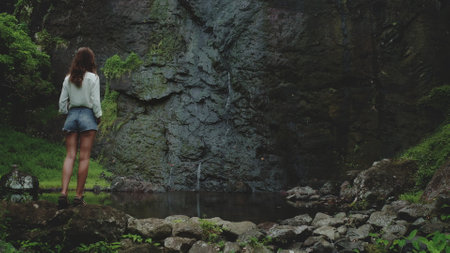 Young woman is enjoying the view of a waterfall cascading down a rocky cliff in a lush tropical forest. The serene atmosphere and the beauty of nature create a sense of peace and tranquilityの写真素材