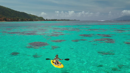 Paddling a yellow kayak in turquoise waters, a woman enjoys a sunny day in a tropical lagoon, surrounded by coral reefs and fluffy cloudsの写真素材