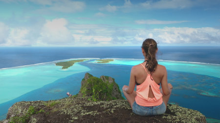 Young woman practices yoga and meditation on a clifftop overlooking the beautiful blue lagoon and motu of maupiti island in french polynesiaの写真素材