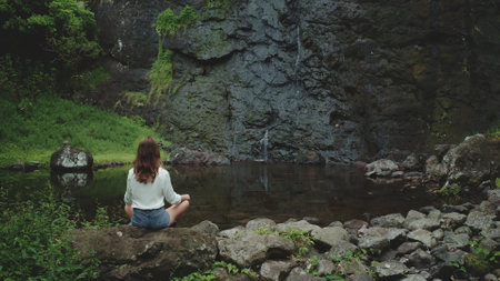 Woman practicing yoga near a small waterfall in a wild tropical jungle. She is enjoying a peaceful moment of mindfulness and connection with natureの写真素材