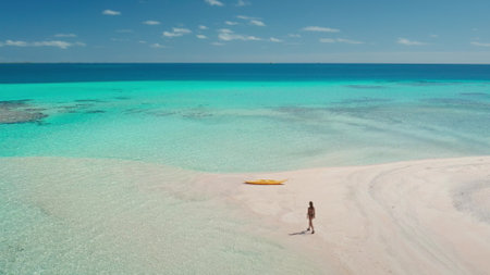 Aerial drone view of a woman walking on a pristine white sand beach towards crystal clear turquoise water with a yellow kayak, creating a sense of wanderlust and tropical paradiseの写真素材