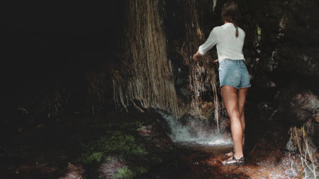 Tourist woman stands near waterfall in tropical jungle forest. Girl picks up water to wash face in hot day. Happy female adult enjoying the water drops. Great wild nature, tourism, travel, holidaysの写真素材