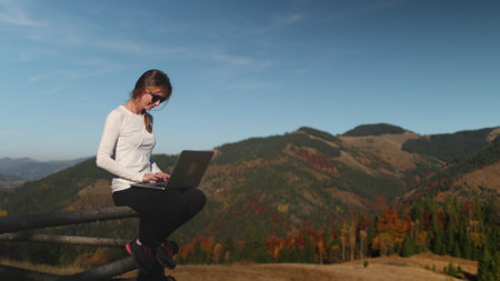A woman is seated comfortably on a rustic wooden fence, engrossed in her laptop screen. Her fingers fly across the keyboard as she works, showcasing the modern blend of nature and technology.の写真素材