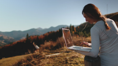 A confident woman stands atop a hill, her eyes fixed on the laptop screen in her hands. She seems both connected to the digital world and in tune with the natural beauty surrounding her.の写真素材