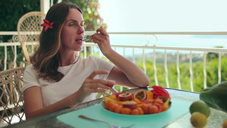 A woman sit wooden table, with a colorful plate of assorted fruits. Reaching for a piece of fruit, ready to enjoy a healthy snack, showcasing the freshness of the exotic fruits.の写真素材
