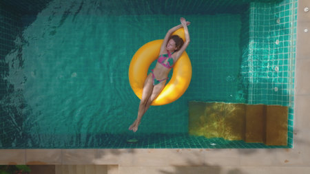 Woman relaxes in a luxurious pool on a sunny day, floating on a yellow inflatable ring, embodying pure bliss and tranquilityの写真素材