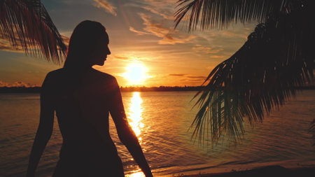 Silhouette of a young woman enjoying a colorful sunset on a tropical beach, with palm trees creating a frame around herの写真素材