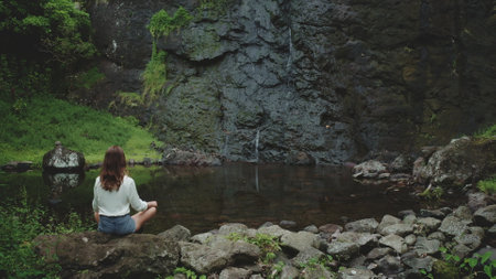 A woman sits meditating on a rock near a waterfall in French Polynesia. The lush green foliage and crystal clear water create a serene and tranquil atmosphere.の写真素材