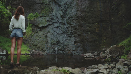 A woman stands on rock waterfall in French Polynesia. The tropical waterfall flows in a small pool, surrounded by lush greenery and volcanic rock. Peace and tranquility, showcasing nature beautyの写真素材