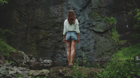 A young woman in a white shirt and denim shorts stands on a rocky outcrop near a waterfall in a lush, tropical forest in French Polynesia. The waterfall cascades down a dark, mossy rock face.の写真素材