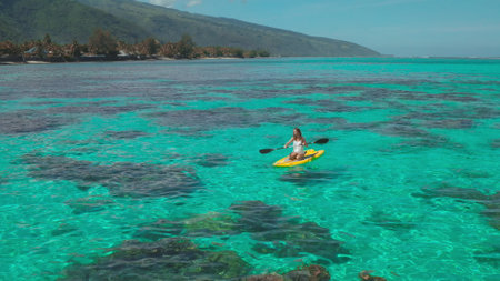 Aerial view of a woman kayaking over a coral reef in turquoise water near a tropical island. She is enjoying a sunny day in paradiseの写真素材