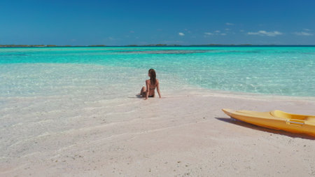 Young woman enjoys the sun and crystal clear water while sitting on a pristine tropical beach with a yellow kayak in the foregroundの写真素材
