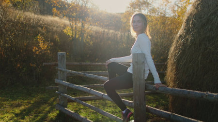 Woman relax outdoor rural village countryside, sitting on wooden fence near a stack of hay. Caucasian female enjoy weekend holiday vacation. Autumn beautiful nature sunset landscape. Slow motionの写真素材