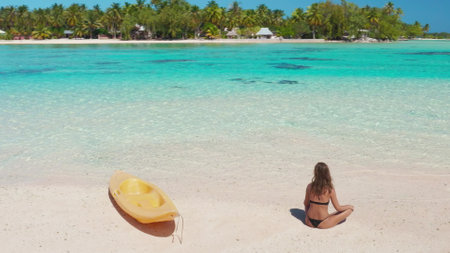 Aerial view of a young woman sitting on the sand of a pristine tropical beach, with a yellow kayak next to her and crystal clear turquoise water in the backgroundの写真素材