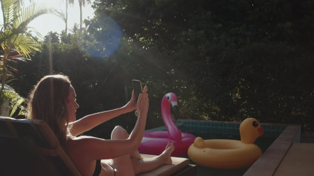 Beautiful girl relax on tropical villa takes selfie with her phone while enjoy sunny day lying near pool in Thailand resort. Girl relax outdoor travel summer holiday vacation in private exotic resortの写真素材