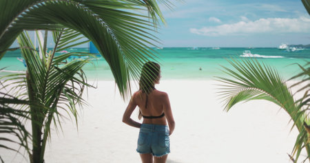 Young woman is taking in the idyllic scenery of a tropical beach, with white sand, turquoise water, and palm trees swaying in the breezeの写真素材