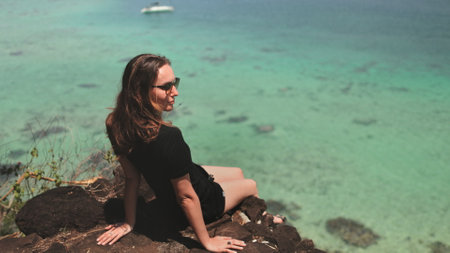 Young tourist is enjoying the view of the ocean from a cliff on a sunny day. The water is a beautiful turquoise color and the sun is shining brightlyの写真素材