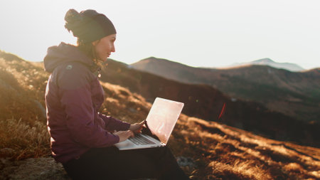 Autumn sunset mountain woman remote working on laptop. Tourist girl typing on notebook sitting on orange grass meadow in soft sun light. Hiking, trekking, outdoor lifestyle travel on holiday vacationの写真素材