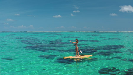 Young woman is paddling on a yellow paddle board in turquoise tropical water, enjoying the sunny weather on a vacation in an exotic countryの写真素材