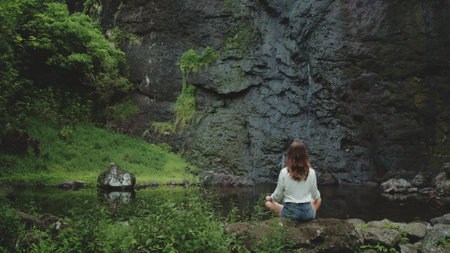 Young woman sits cross-legged on a rock, meditating in front of a lush waterfall in French Polynesia. The tropical landscape is serene and peaceful, with small pool of water. Back view, dark toningの写真素材