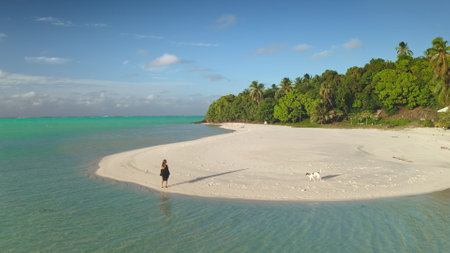 Woman is enjoying a sunny day with her dog on a pristine white sand beach, with lush green tropical vegetation and turquoise waterの写真素材