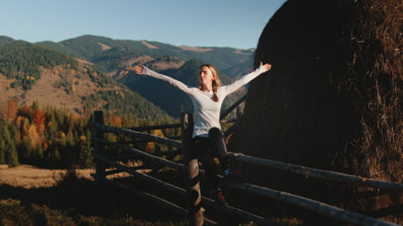 Young woman sits on a wooden fence in a stunning mountain landscape, embracing freedom in lush forests, autumn colors, and a sunny sky. Enjoying the peaceful carpathian mountains of ukraineの写真素材